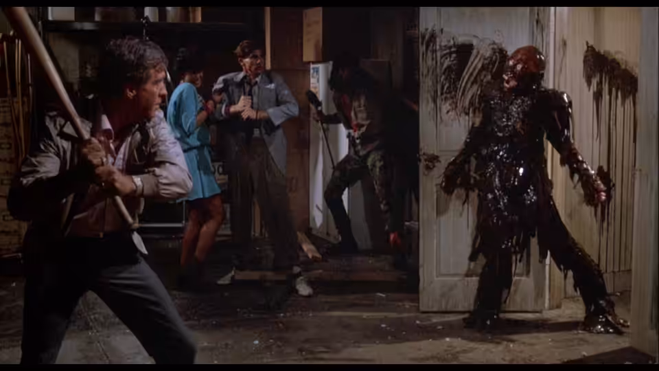 Burt braces with a weapon as friends confront a bloodied corpse in a doorway