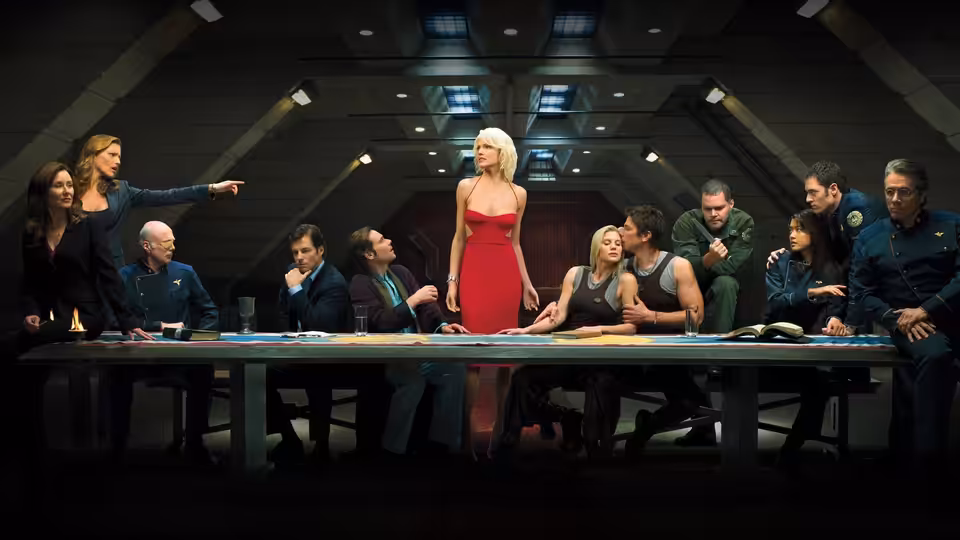 Number Six (Tricia Helfer) in red dress stands at head of long table