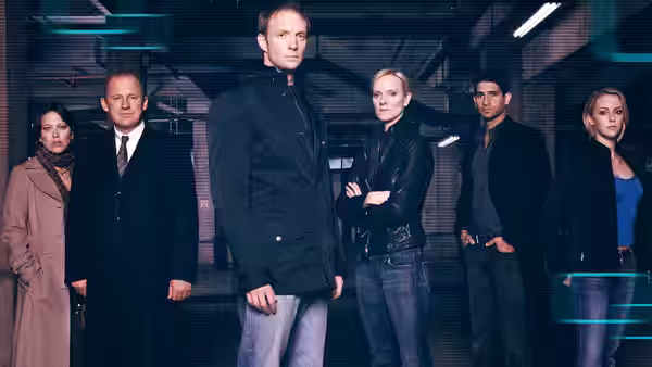 Harry Pearce (Peter Firth) stands with five team members in an industrial office space