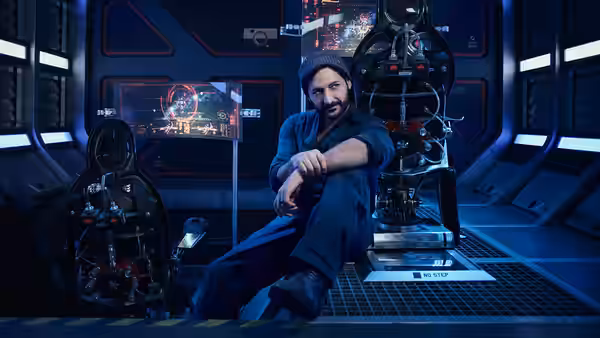 A bearded man in a beanie lounges in a high-tech control room