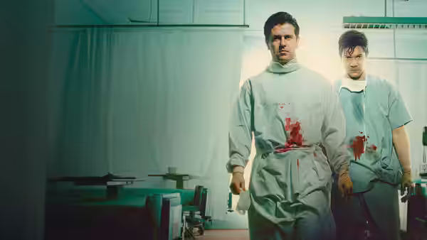 Two men in bloodied surgical gowns walk through hospital ward