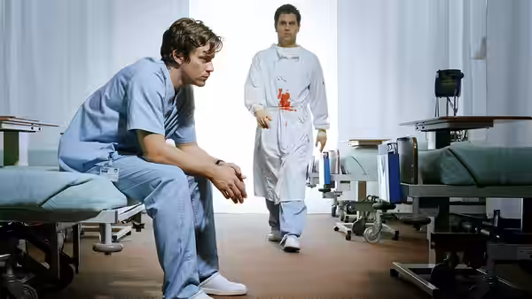 Man in blue scrubs sits on hospital bed as blood-stained colleague stands nearby