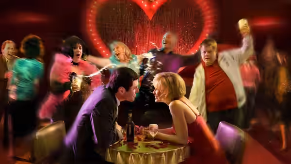 A couple at a table shares a drink while a group dances in a red-lit club