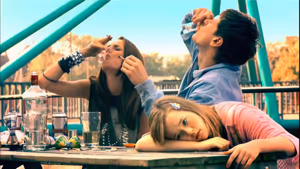 Three teenagers at an outdoor table with one drinking and one resting head