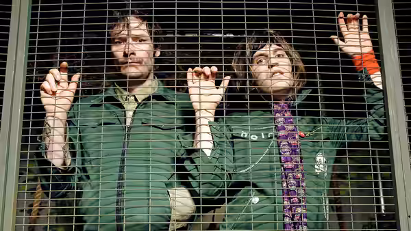 Howard Moon (Julian Barratt) and Vince Noir (Noel Fielding) behind a metal fence with hands raised