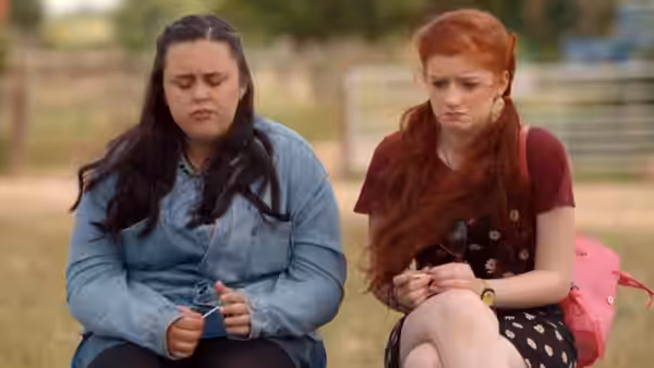 Rachel 'Rae' Earl (Sharon Rooney) sits beside a red-haired friend, holding a small object