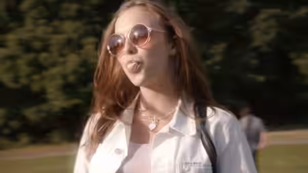 A young woman in sunglasses walks through a park, making a face