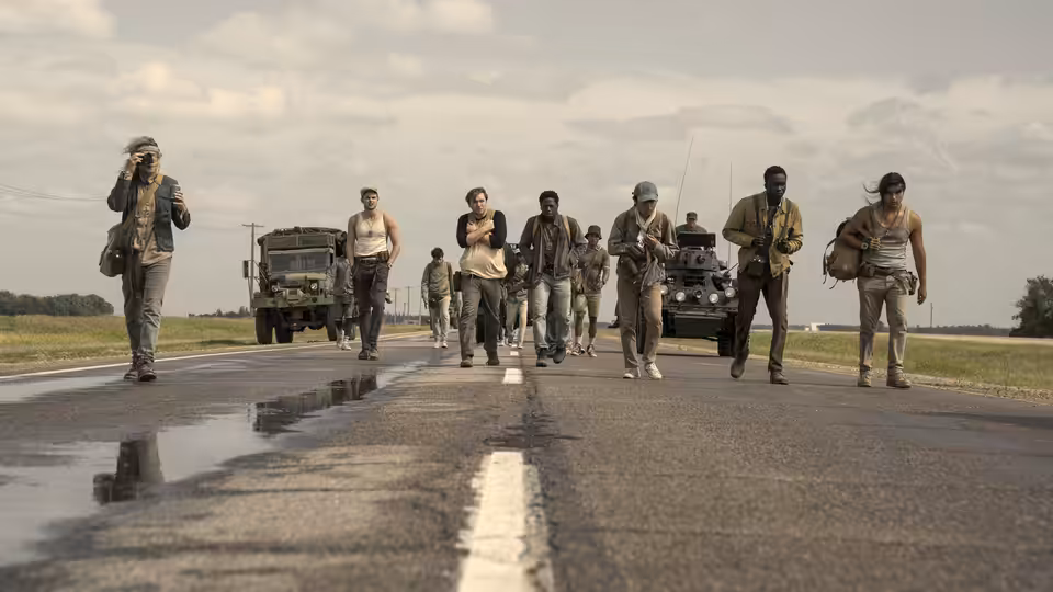 A group of young men walking along a paved road with military trucks following