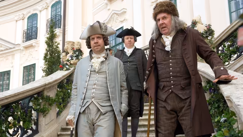 John Adams (Paul Giamatti) and Benjamin Franklin (Tom Wilkinson) descending stairs outside a palace