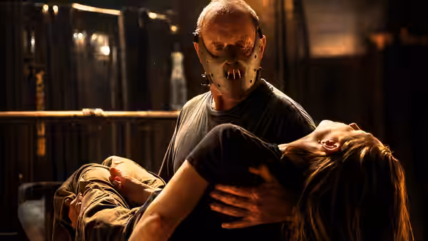 Hannibal Lecter (Anthony Hopkins), wearing a muzzle, carries an unconscious woman through a dim room