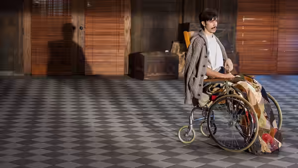 Wallace Bryton (Justin Long) stands behind a wheelchair piled with blankets in a tiled room
