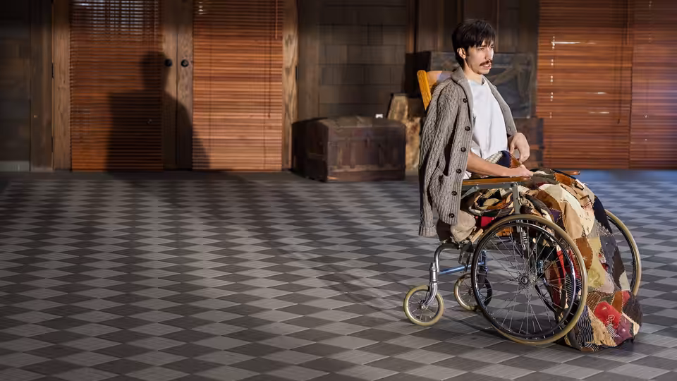 Wallace Bryton (Justin Long) stands behind a wheelchair piled with blankets in a tiled room