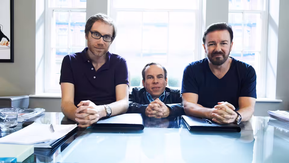 Stephen Merchant (Stephen Merchant), Warwick Davis (Warwick Davis) and Ricky Gervais (Ricky Gervais) seated at a glass desk