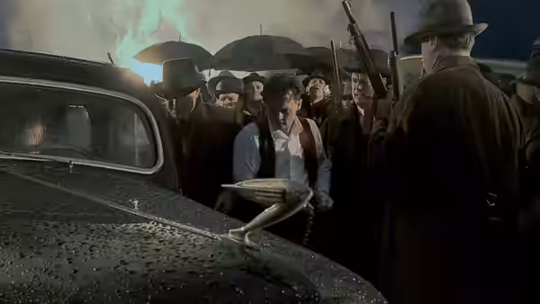 A man holding a hat stands beside a car as armed men gather around him