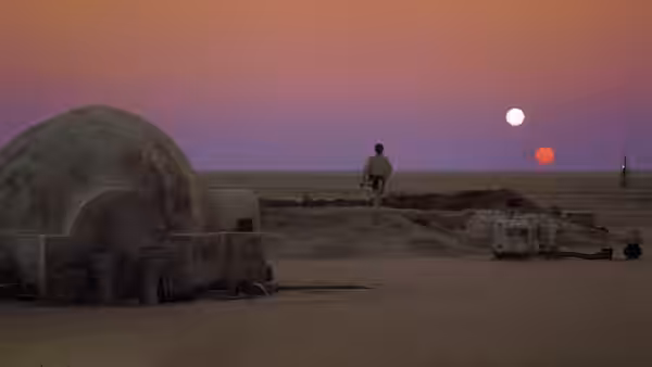 a figure in brown walks past a domed structure across sandy ground under two suns