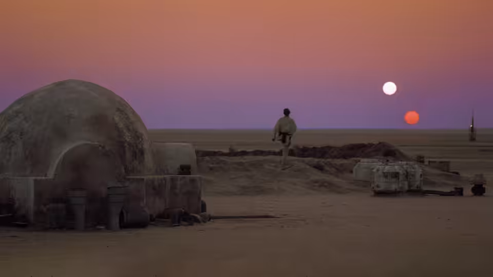 a figure in brown walks past a domed structure across sandy ground under two suns