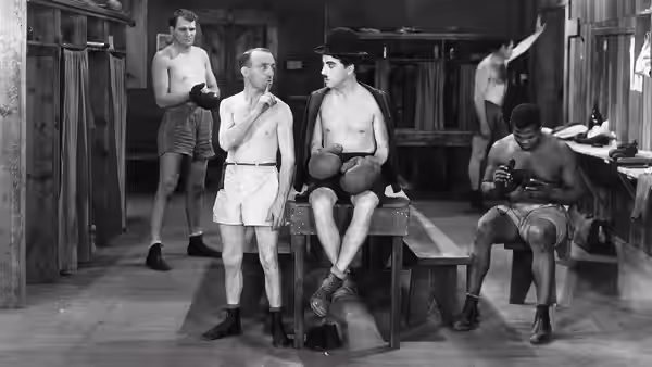 The Tramp (Charlie Chaplin) sits on a bench in a boxing locker room