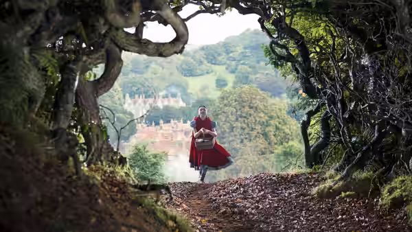 Little Red Riding Hood (Lilla Crawford) walks through a wooded arch toward a distant castle