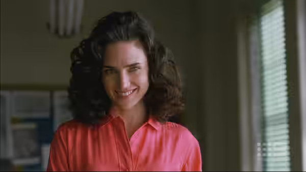 Alicia Nash (Jennifer Connelly) smiling in a pink collared shirt indoors