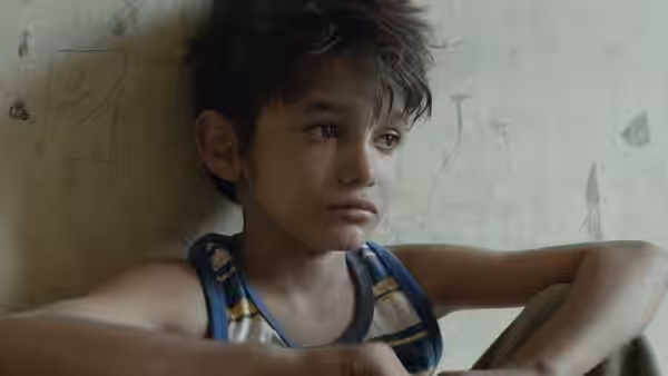 A shirtless boy leans against a wall with a sad expression