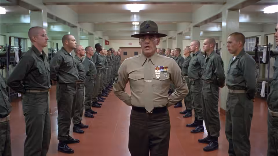 Gny. Sgt. Hartman (R. Lee Ermey) stands hands on hips addressing recruits in formation indoors