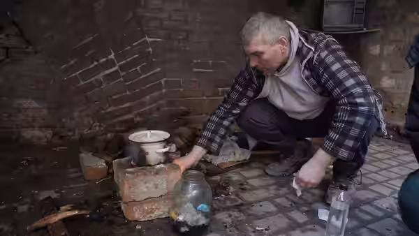 Man crouching beside a makeshift brick stove in a destroyed room adjusting a pot