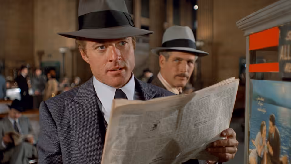 Man in fedora reads newspaper in busy station as another watches