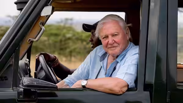 David Attenborough seated in a green vehicle beside a driver in open country
