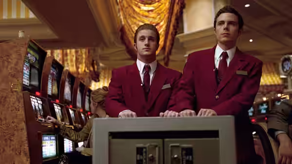 Two men in red uniforms standing behind slot machines in a casino
