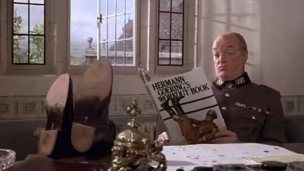 A uniformed officer reads a book at a desk in a room with tall windows