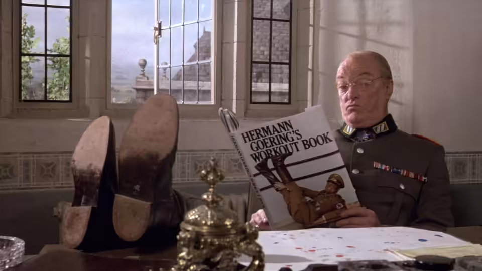 A uniformed officer reads a book at a desk in a room with tall windows