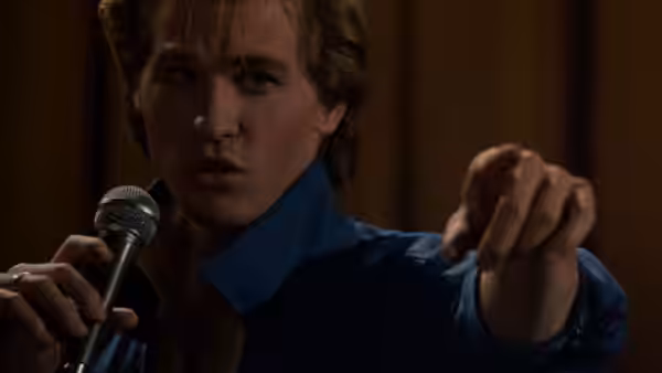 Nick Rivers (Val Kilmer) sings into a microphone and points toward the audience