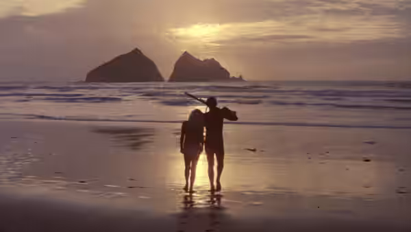 A couple walk arm in arm along the wet sand toward the setting sun