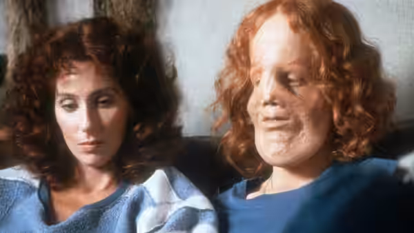 Rusty Dennis (Cher) sits beside Rocky Dennis (Eric Stoltz) indoors