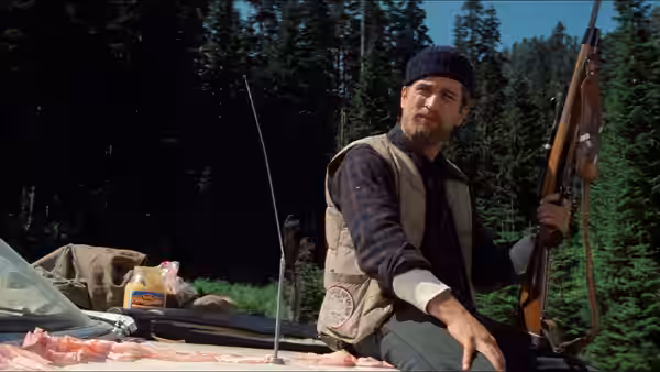 Bearded man in vest holding rifle beside vehicle in forest clearing