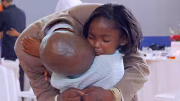 A bald man bends down, embracing a young girl in a bright room