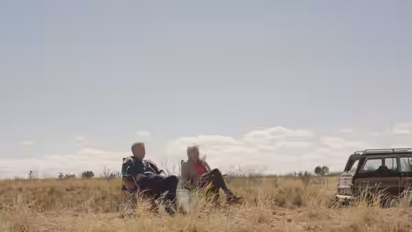 Will Ferrell and Harper Steele sit in dry grass beside their parked SUV