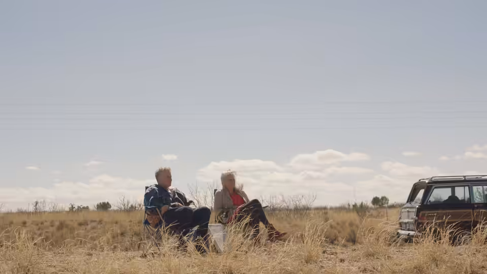Will Ferrell and Harper Steele sit in dry grass beside their parked SUV