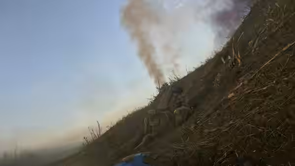 A soldier crouches on a wooded slope beneath rising smoke