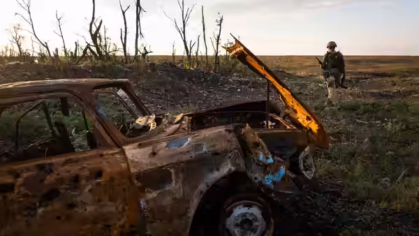 A soldier crosses a field beside a burned-out car