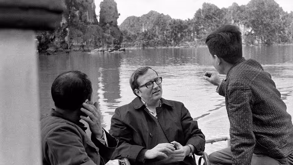 Three men sit beside a lake as one gestures toward the water