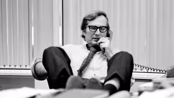 A bespectacled man sits in an office chair, speaking on a telephone