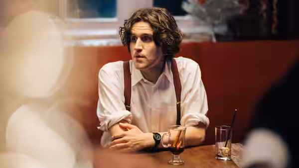 Colin (Harry Melling) sits at a bar table, listening with his arms folded