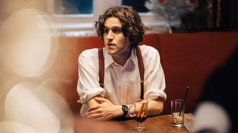 Colin (Harry Melling) sits at a bar table, listening with his arms folded