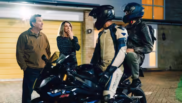 Two motorcycle riders in helmets sit on a bike as a man and woman watch