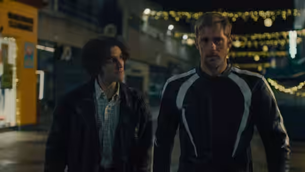 Colin (Harry Melling) walks beside Ray (Alexander Skarsgård) along a festoon-lit street