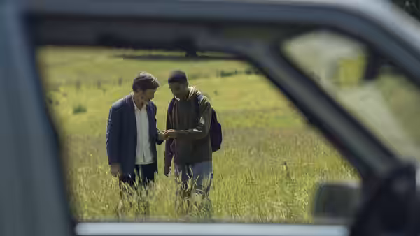 Steve and a young man examine something in a grassy field, seen through a car window