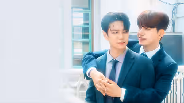 A suited man stands behind another, holding his hands in a bright room