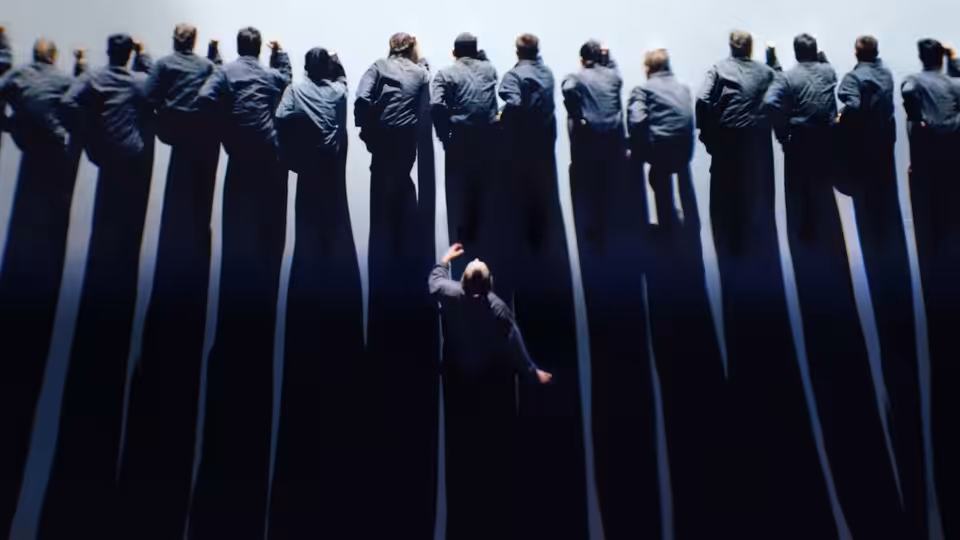 A man reaches toward a line of dancers standing above him against a dark backdrop
