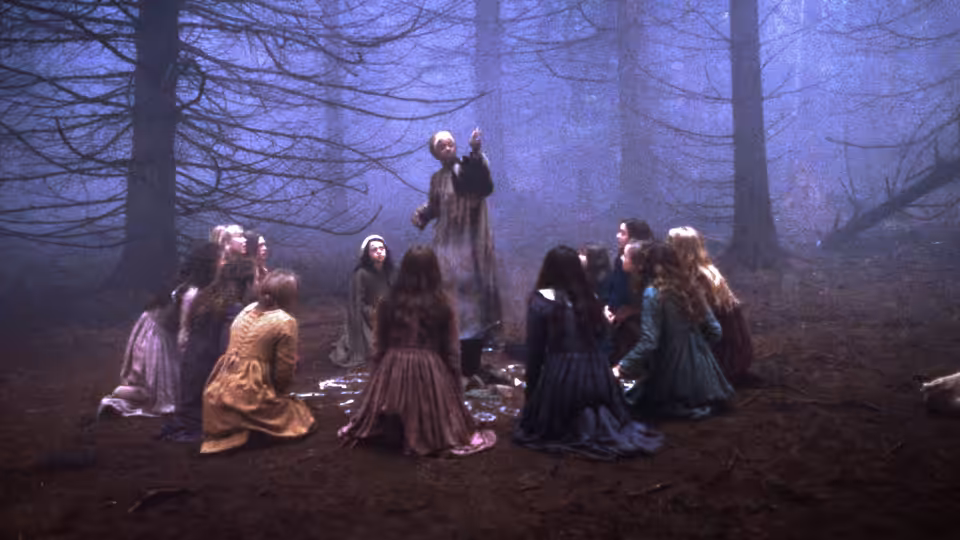 A group of young women gather around a standing figure in a misty forest